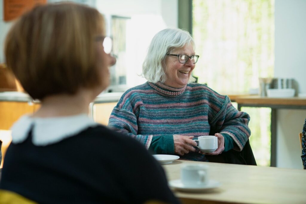 Older woman wearing glasses and a striped sweater smiling while holding a cup of tea at a table indoors.